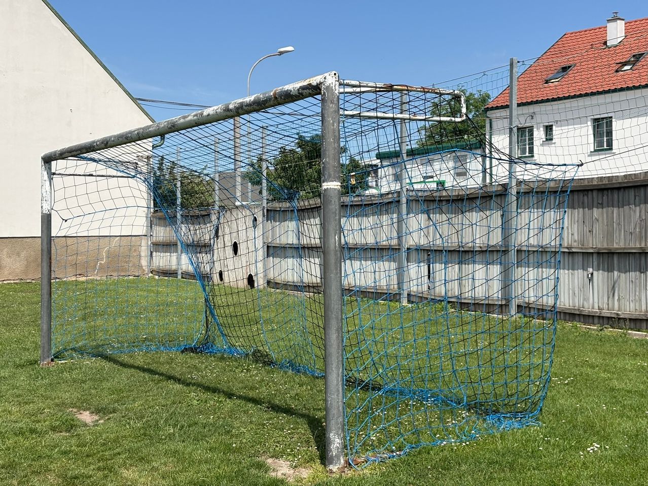 A rusty metal soccer goal with blue nets is set up on a grassy field, surrounded by a white fence and a house.