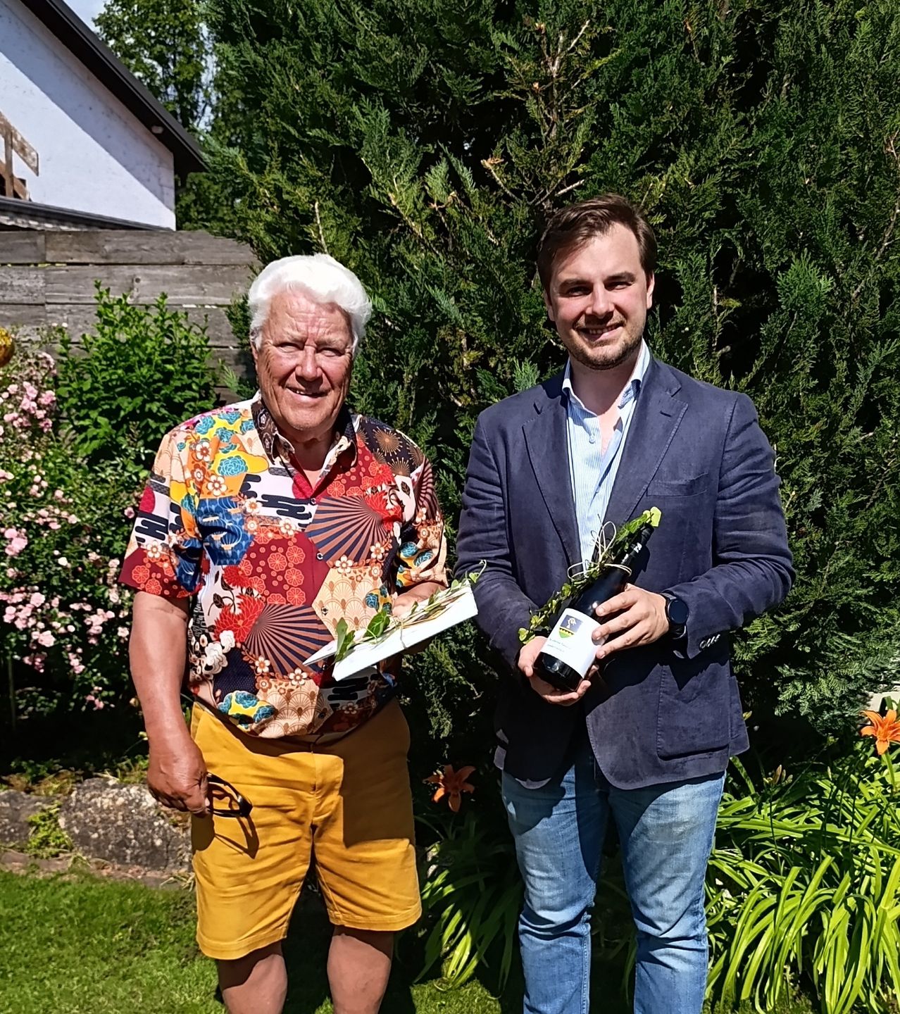 Zwei Männer stehen in einem Garten mit einem Blumenstrauß und einer Flasche. Der ältere Mann trägt ein farbenfrohes Hemd und gelbe Shorts. Der jüngere Mann hält eine Flasche und eine Pflanze.