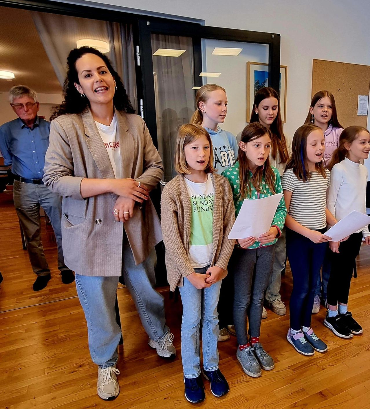 Eine Gruppe junger Mädchen, die Papiere in der Hand halten, steht in einem Raum vor einer Frau. Dahinter steht ein Mann und eine Anschlagtafel.