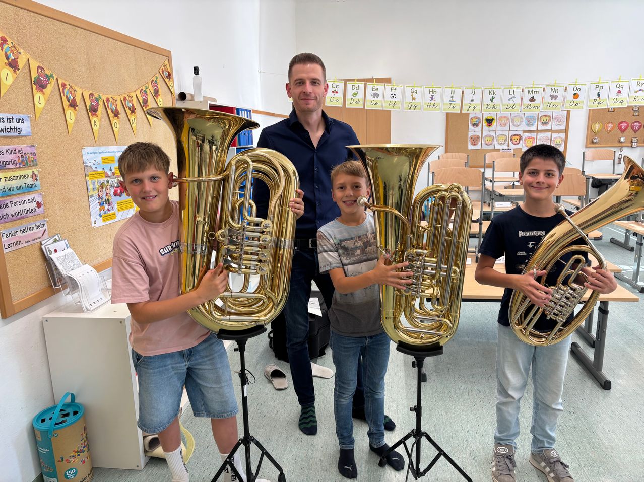Drei Jungen halten große Blechblasinstrumente in einem Klassenzimmer. Der Junge auf der linken Seite trägt ein rosa Hemd, der Junge in der Mitte trägt ein graues Hemd und der Junge auf der rechten Seite trägt ein blaues Hemd. Hinter ihnen steht ein Mann in einem blauen Hemd mit lächelndem Gesicht.