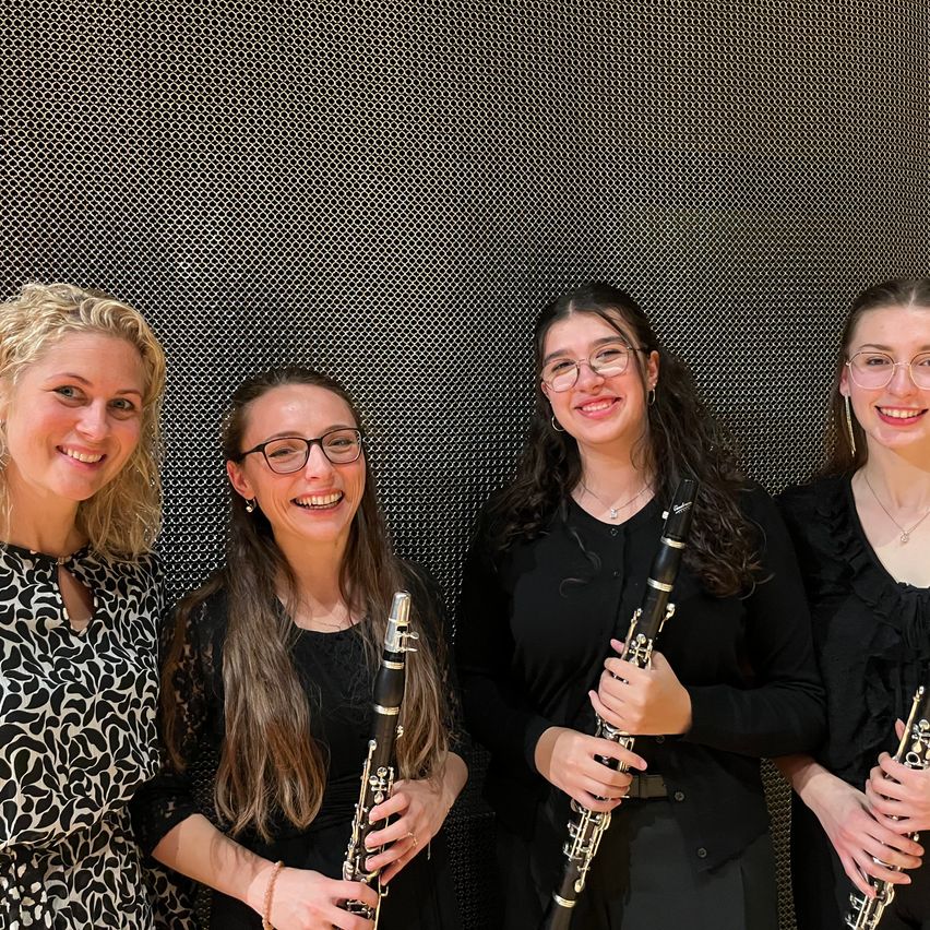 Vier Frauen stehen eng beieinander und lächeln. Drei von ihnen halten Klarinetten. Sie sind in Schwarz gekleidet. Der Hintergrund ist eine dunkle strukturierte Wand.