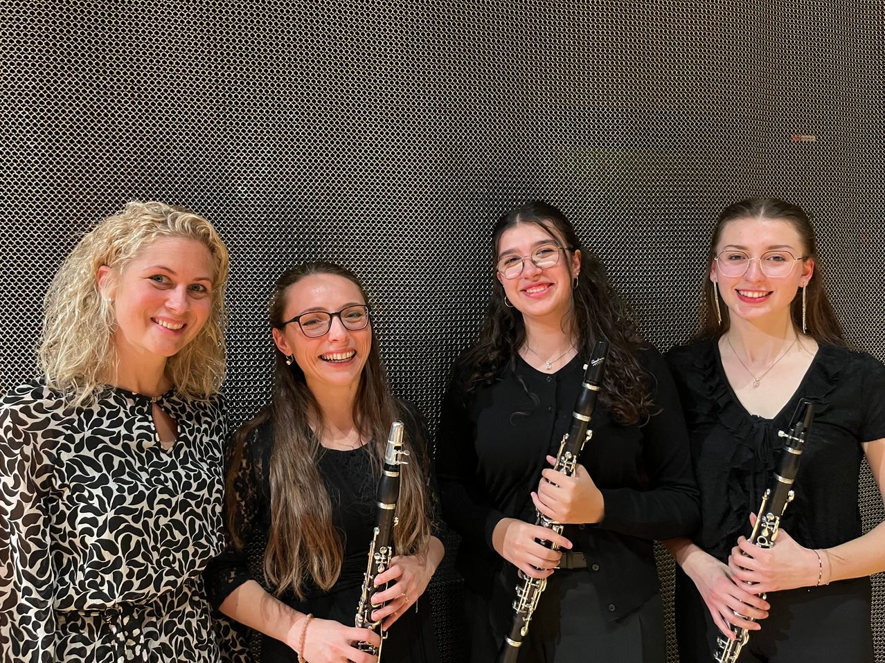 Vier Frauen stehen eng beieinander und lächeln. Drei von ihnen halten Klarinetten. Sie sind in Schwarz gekleidet. Der Hintergrund ist eine dunkle strukturierte Wand.