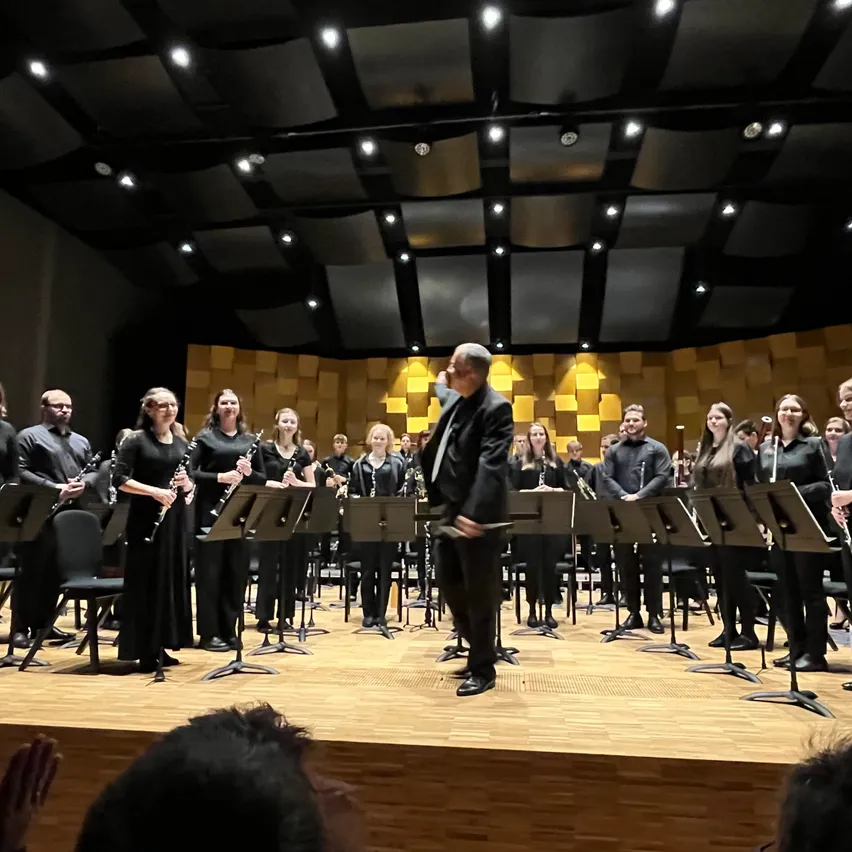 Ein Klarinettenensemble spielt auf der Bühne mit einem Dirigenten. Die Musiker sind in Schwarz gekleidet, und die Bühne ist von Deckenleuchten beleuchtet.
