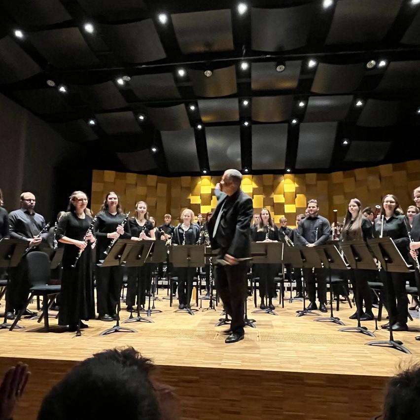 Ein Klarinettenensemble spielt auf der Bühne mit einem Dirigenten. Die Musiker sind in Schwarz gekleidet, und die Bühne ist von Deckenleuchten beleuchtet.