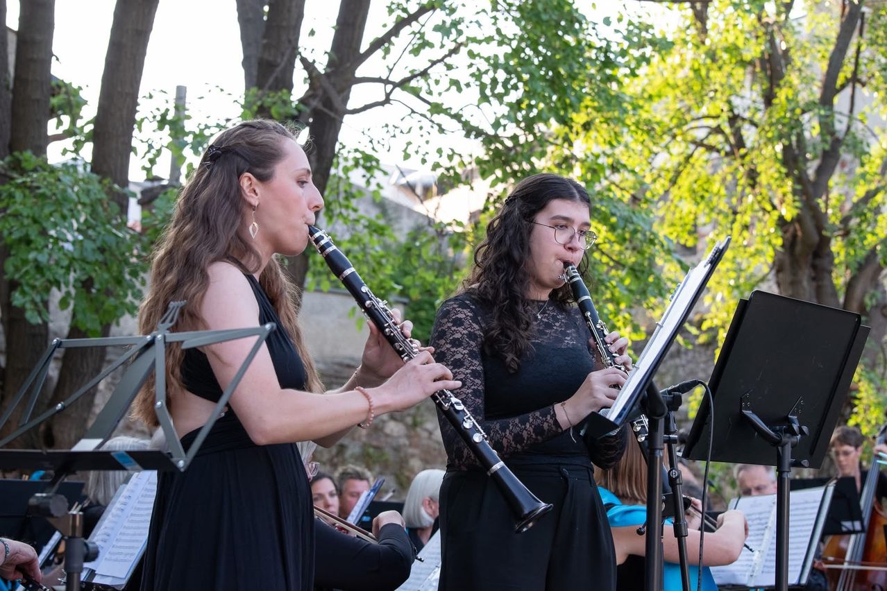 Zwei Frauen spielen Klarinette und stehen vor einem Mikrofon. Dahinter befinden sich Bäume und andere Musiker, die auftreten. Der Ort scheint im Freien zu sein.