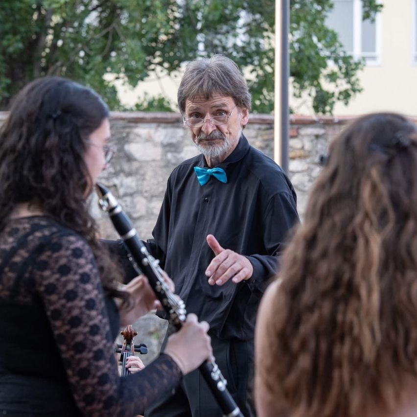 Ein älterer Mann mit Brille und einer blauen Schleife gibt einer jungen Frau, die eine Klarinette hält, Anweisungen. Eine andere Frau hört hinter ihr zu. Sie befinden sich in einer Außenumgebung mit einer Steinmauer und Bäumen im Hintergrund.