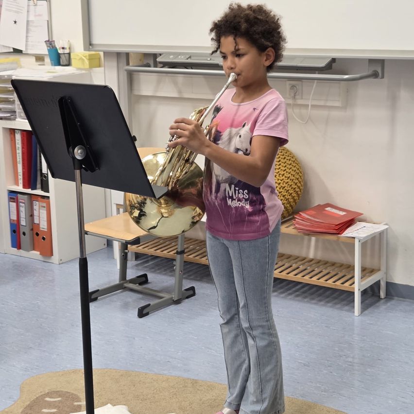 Ein junges Mädchen steht in einem Klassenzimmer und spielt eine Trompete, während sie Notenblätter auf einem Ständer betrachtet. Hinter ihr sind eine Tafel, Regale und ein Tisch mit Büchern zu sehen.