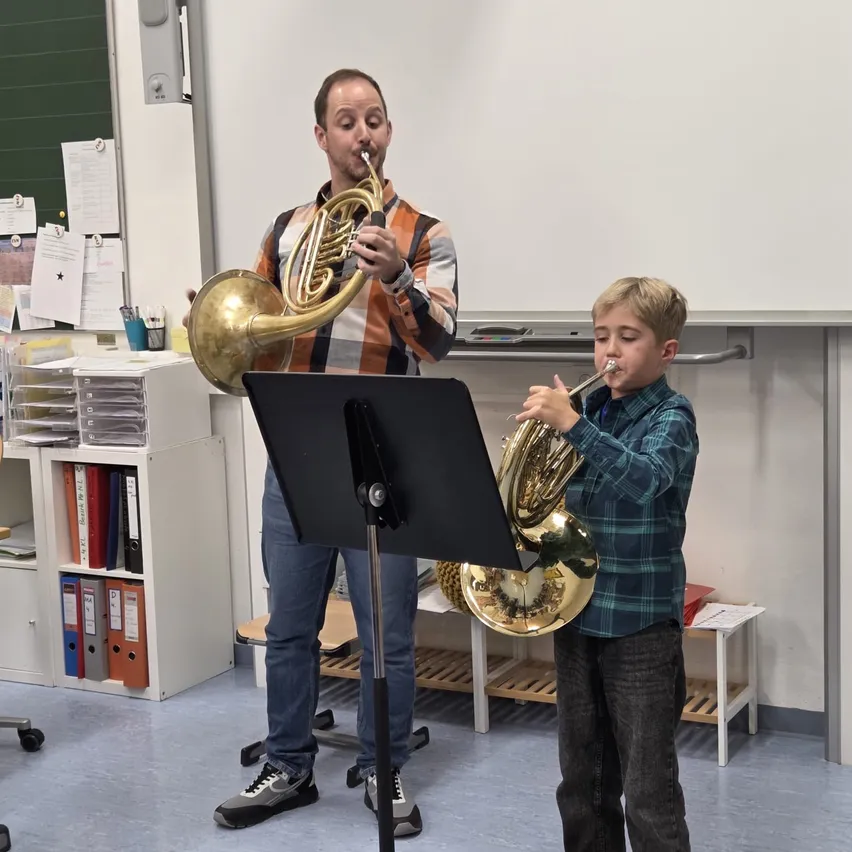 Ein Mann und ein Junge stehen in einem Klassenzimmer und spielen Blechblasinstrumente. Der Mann spielt Trompete und der Junge Tuba. Sie stehen vor einer Tafel und einem Notenständer.