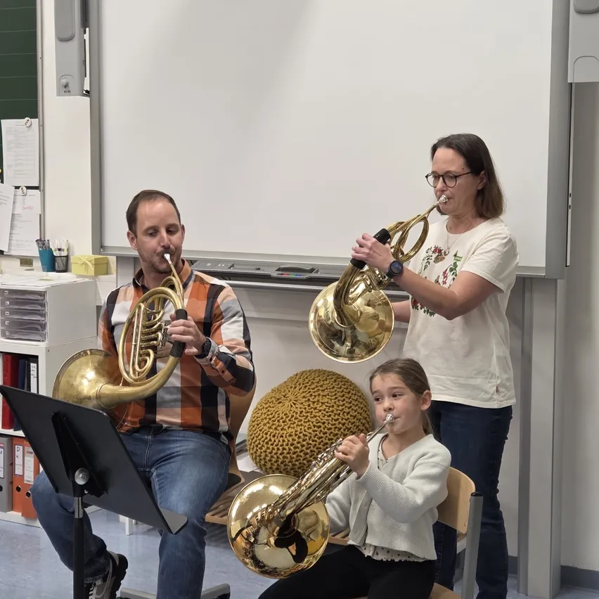 Drei Personen spielen Trompeten in einem Klassenzimmer. Ein Mann sitzt auf einem Stuhl und spielt eine Trompete, und eine Frau steht und spielt eine weitere Trompete. Ein Mädchen sitzt auf einem Stuhl vor ihnen und spielt eine Trompete. Hinter ihnen steht eine Tafel.