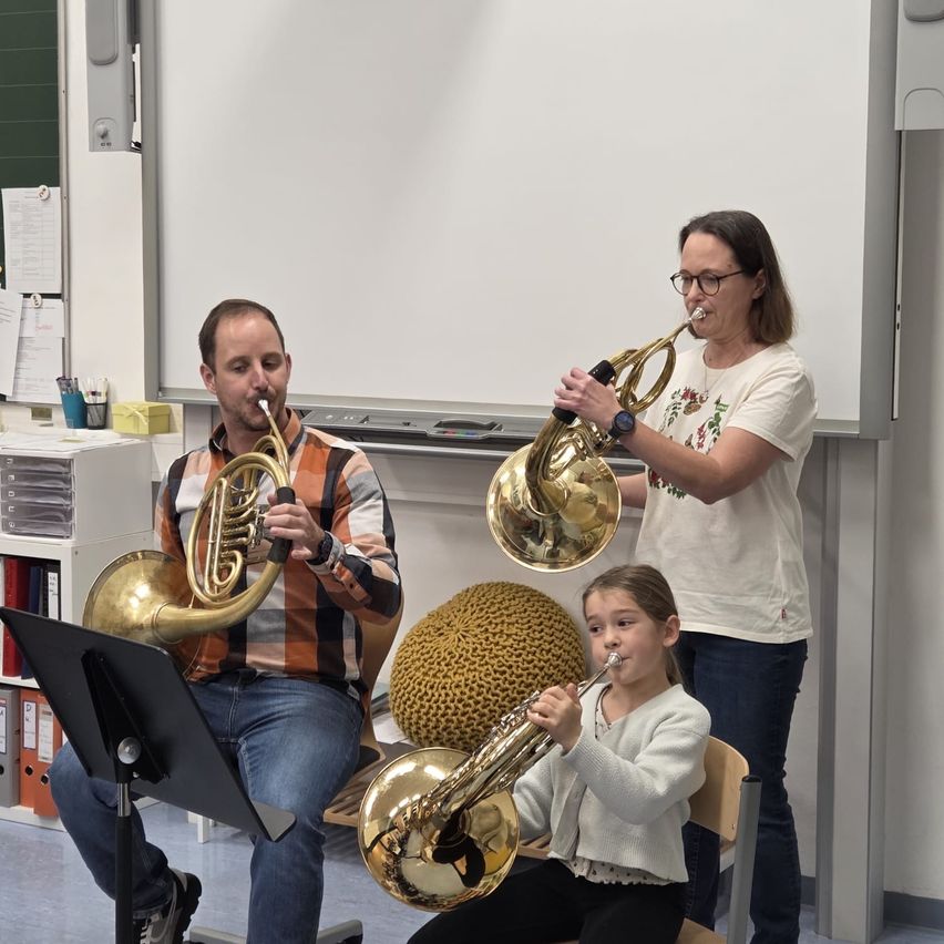 Drei Personen spielen Trompeten in einem Klassenzimmer. Ein Mann sitzt auf einem Stuhl und spielt eine Trompete, und eine Frau steht und spielt eine weitere Trompete. Ein Mädchen sitzt auf einem Stuhl vor ihnen und spielt eine Trompete. Hinter ihnen steht eine Tafel.