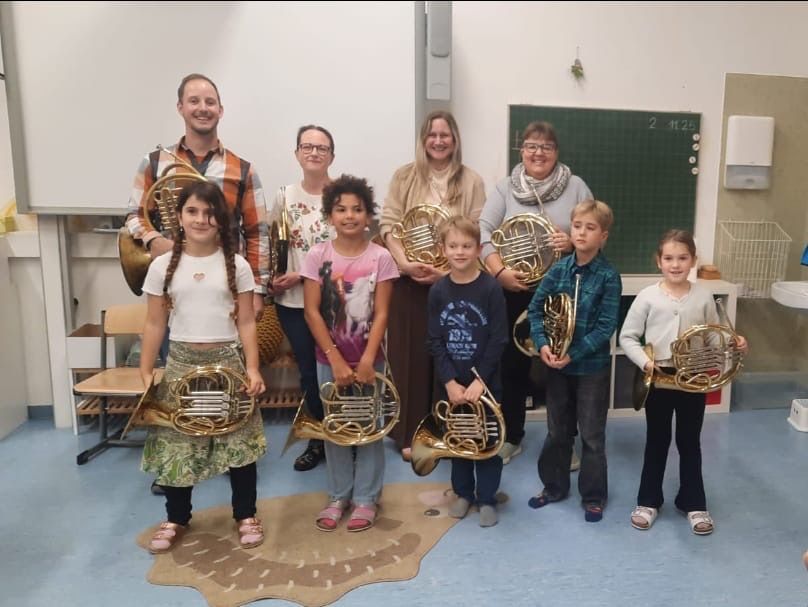 Eine Gruppe von Kindern und zwei Erwachsenen sind in einem Klassenzimmer. Die Kinder halten Blechblasinstrumente und die Erwachsenen lächeln. Das Klassenzimmer hat eine grüne Tafel mit einem Whiteboard und einer Tafel. Es gibt Stühle und einen Schreibtisch im Raum.