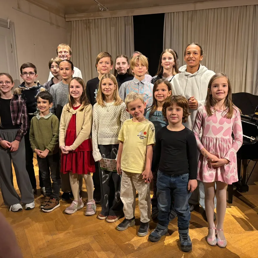 Eine Gruppe von Kindern und einem Mann stehen in einem Raum mit einem Klavier im Hintergrund. Sie alle lächeln und posieren für ein Foto.