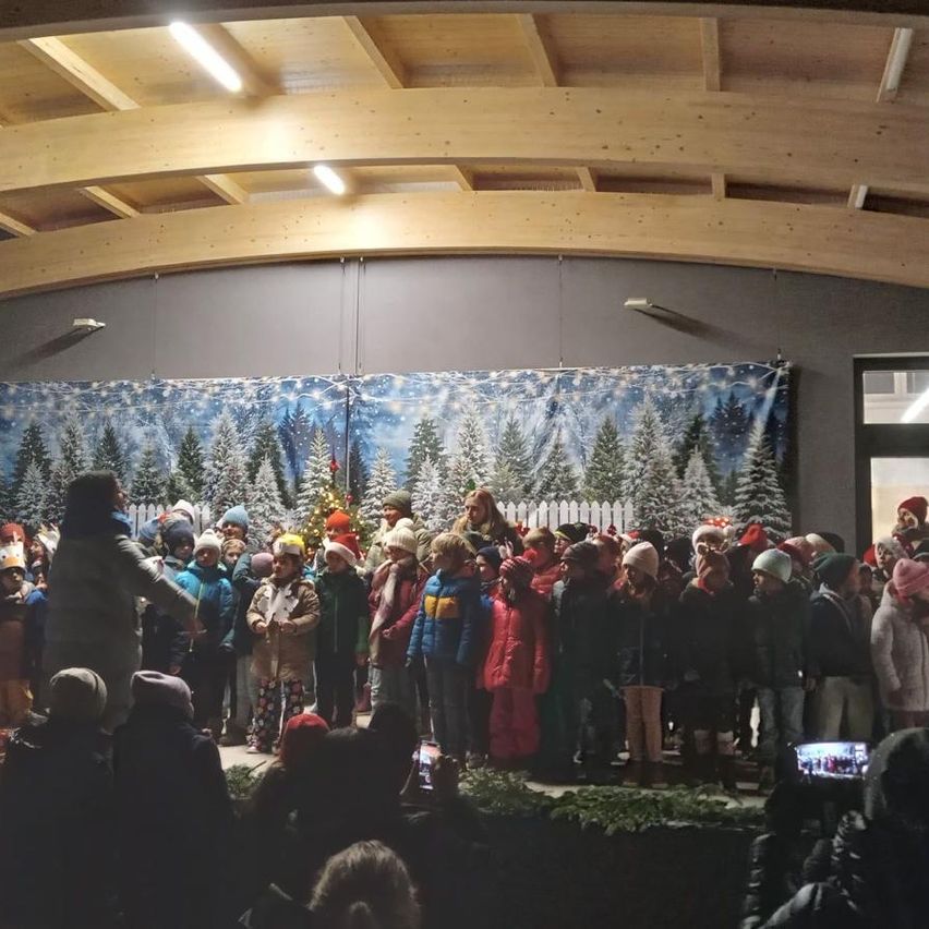 Eine Menschenmenge, hauptsächlich Kinder, versammelt sich in einem Innenraum, möglicherweise für eine Aufführung. Dahinter stehen ein Weihnachtsbaum und eine Kulisse mit Winterthema.