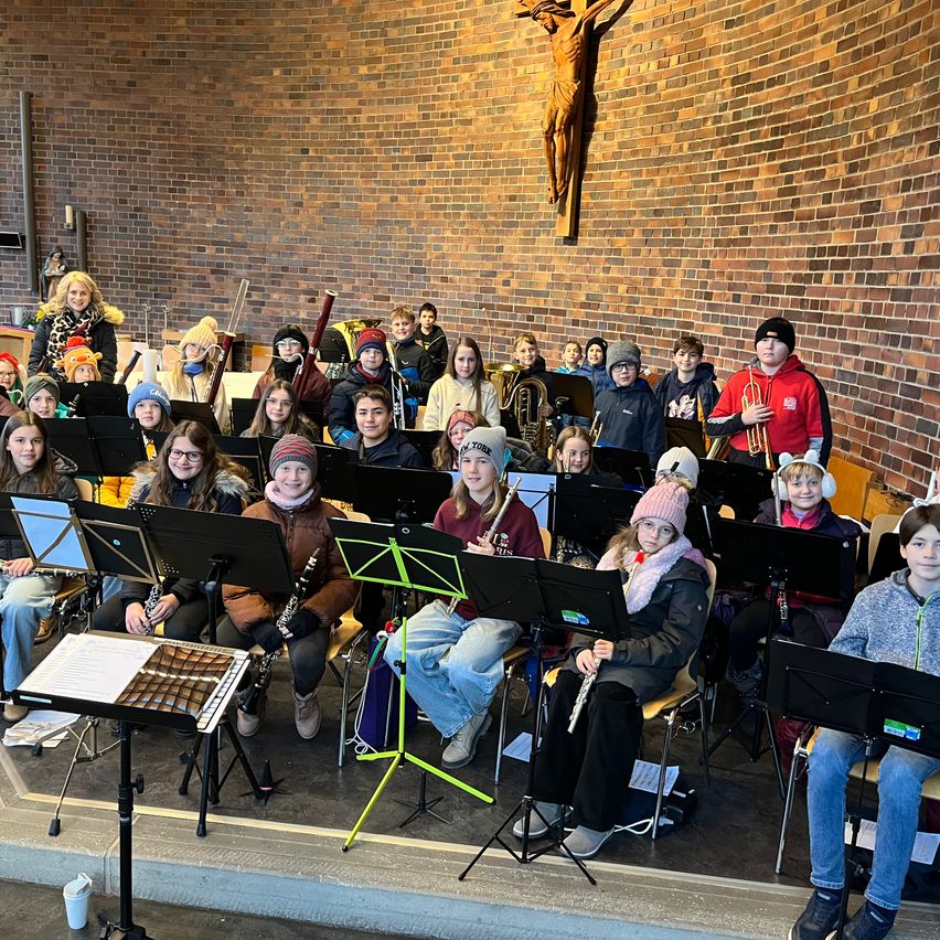 Eine große Gruppe von Schülern, die Winterkleidung tragen, sitzt in einem Raum mit Ziegelwänden. Sie halten verschiedene Musikinstrumente wie Trompeten, Klarinetten und Flöten. Der Raum hat eine Bühne mit Notenständern und ein Kreuz an der Wand.
