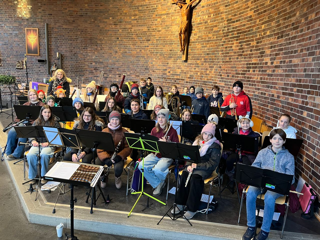 Eine große Gruppe von Schülern, die Winterkleidung tragen, sitzt in einem Raum mit Ziegelwänden. Sie halten verschiedene Musikinstrumente wie Trompeten, Klarinetten und Flöten. Der Raum hat eine Bühne mit Notenständern und ein Kreuz an der Wand.