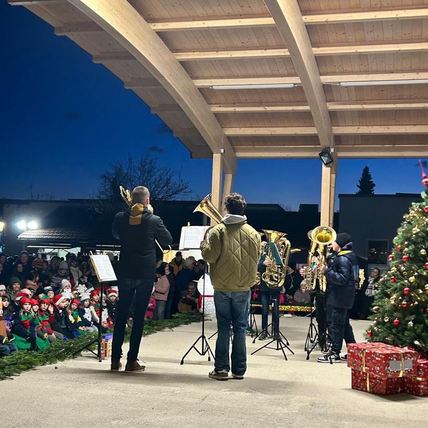 Bei Nacht spielen eine Gruppe von Menschen auf der Bühne mit Musikinstrumenten vor einem Publikum. Ein Weihnachtsbaum ist mit Lichtern und Geschenken geschmückt.