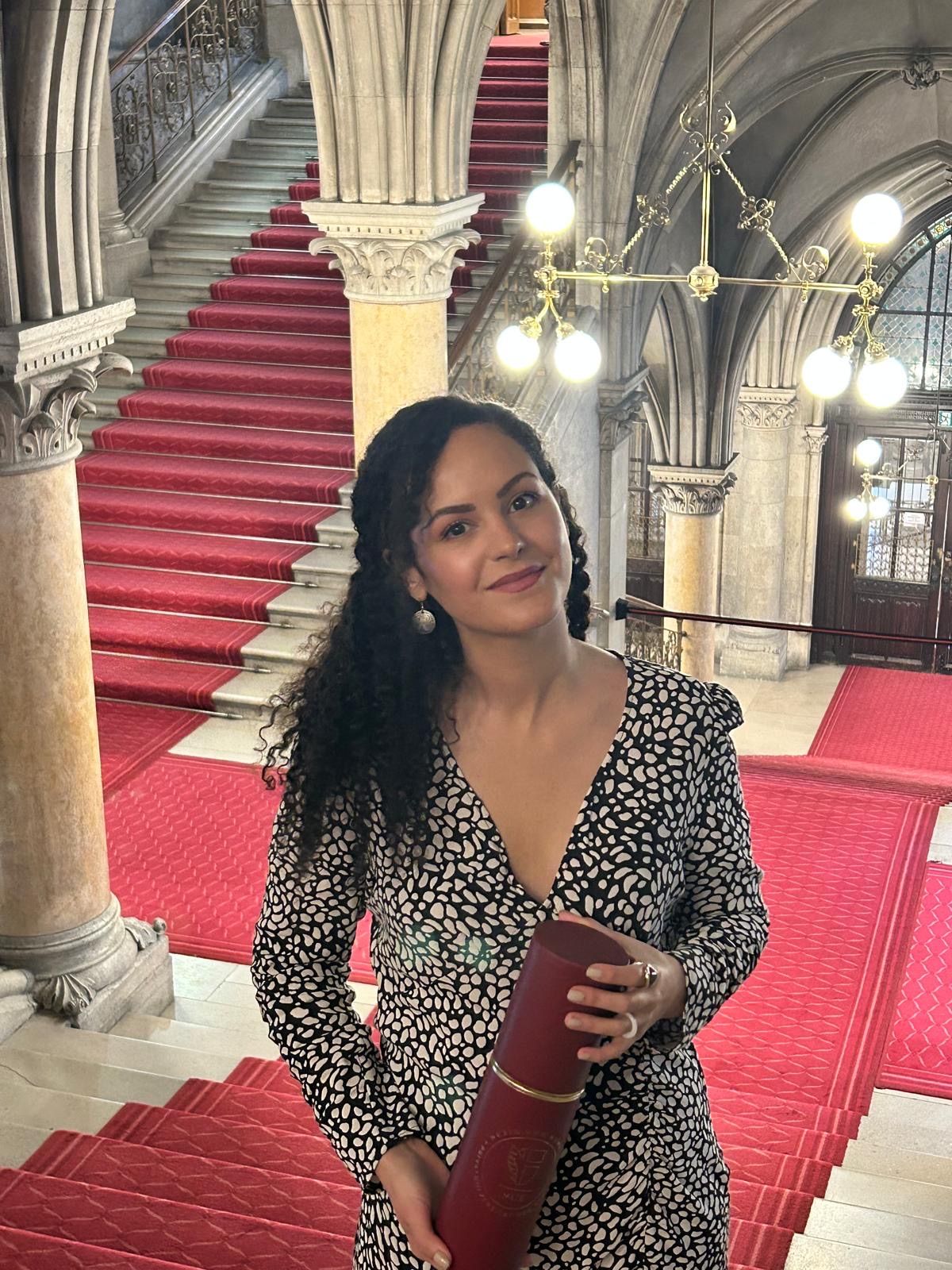 Eine Frau mit lockigem Haar steht auf einem roten Teppich in einem Flur mit Marmorsäulen und Treppen. Sie hält ein rotes Objekt in den Händen.