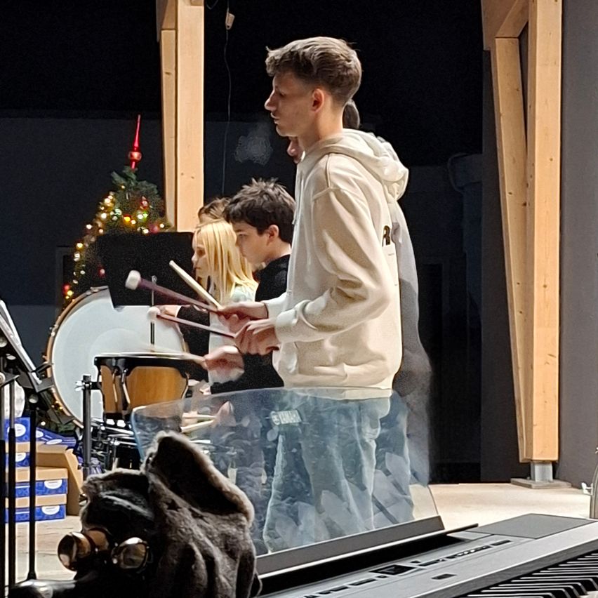 Eine Gruppe junger Musiker spielt mit Trommeln und Becken. Ein Junge in einer weißen Kapuzenjacke spielt die Trommeln, während andere Trommelstöcke halten. Ein Weihnachtsbaum und Kisten sind im Hintergrund.