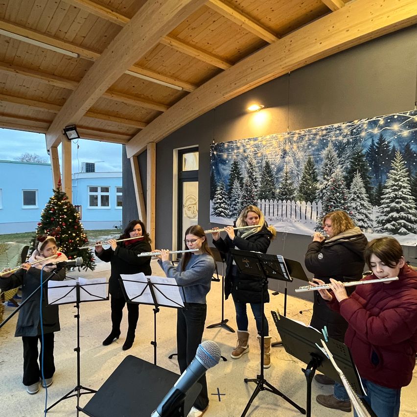 Eine Gruppe von Frauen spielt Flöten in einem Raum mit Holzdecken. Der Raum hat einen Weihnachtsbaum und eine weihnachtliche Wanddekoration. Sie stehen vor Notenständern und Mikrofonen.
