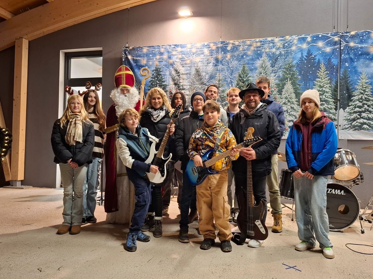 Eine Gruppe von Menschen steht vor einem weihnachtlich gestalteten Hintergrund und posiert für ein Foto. Einige halten Gitarren, während andere dahinter stehen. In der Mitte steht eine als Weihnachtsmann verkleidete Person.