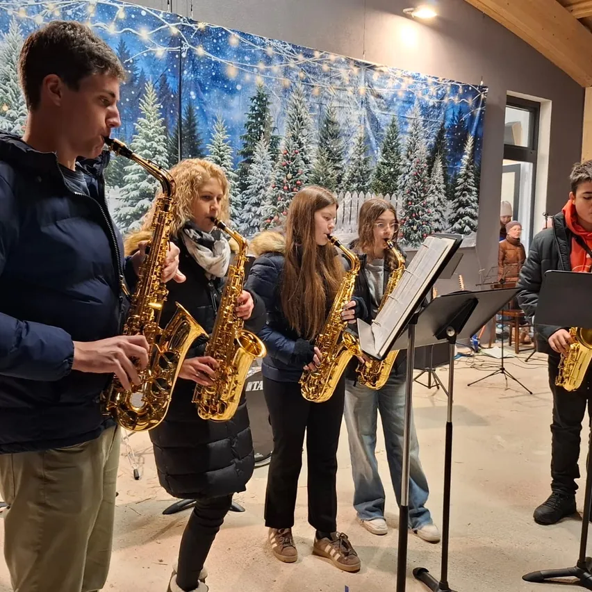Eine Gruppe von Musikern spielt Saxophon in einem Raum mit einer weihnachtlichen Hintergrundkulisse. Zwei Frauen und ein Mann spielen, während eine Frau Musik von einem Notenständer liest.