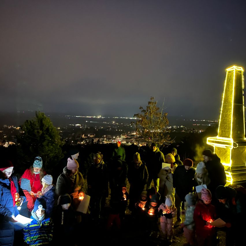Eine Gruppe von Menschen hält Kerzen im Dunkeln. Sie sind vor einem hohen gelben Turm versammelt.