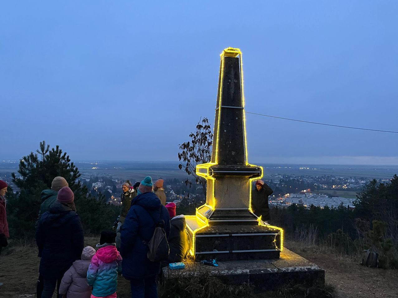 Eine Gruppe von Menschen versammelt sich um einen beleuchteten Obelisken auf einem Hügel bei Dämmerung, mit Stadtlichtern in der Ferne.