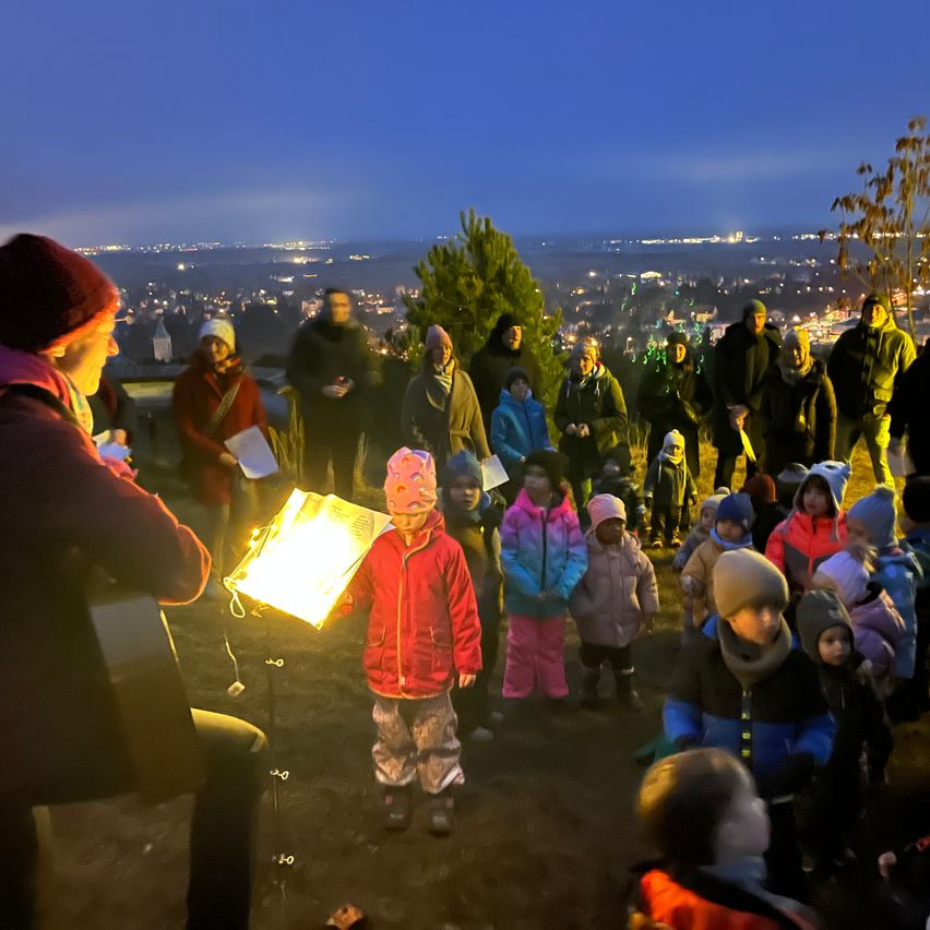 Eine Gruppe von Menschen, darunter Kinder, versammelt sich im Freien bei Dämmerung in einem Kreis. Eine Frau hält eine beleuchtete Papierlaterne und eine Musikpartitur. Die Stadtlichter sind in der Ferne sichtbar.