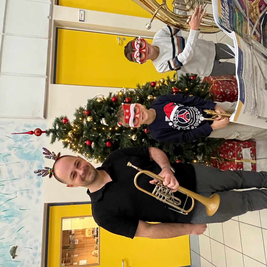 Drei Personen, alle mit festlichen Brillen, stehen vor einem geschmückten Weihnachtsbaum. Der Mann vor ihnen hält eine Trompete, während die beiden dahinter ein Tuba und ein Posaune spielen.