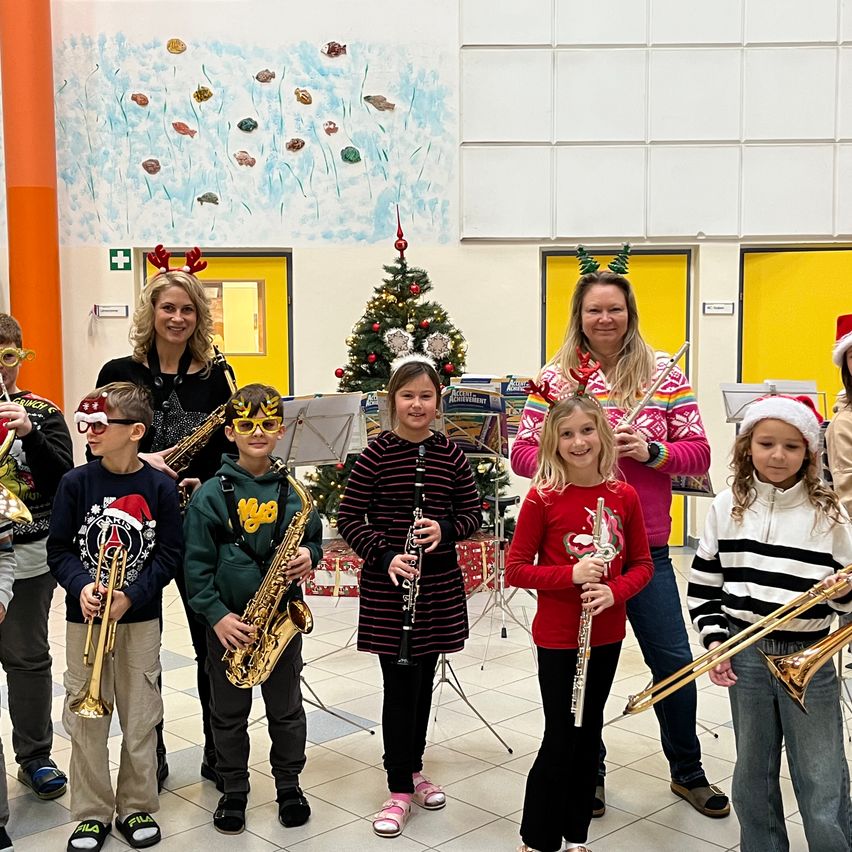 Eine Gruppe von Kindern und zwei Erwachsenen steht in einem Flur. Sie sind in weihnachtlich thematisierten Kleidungsstücken gekleidet und halten Blechblasinstrumente. Ein Weihnachtsbaum und Weihnachtsdekorationen sind im Hintergrund.