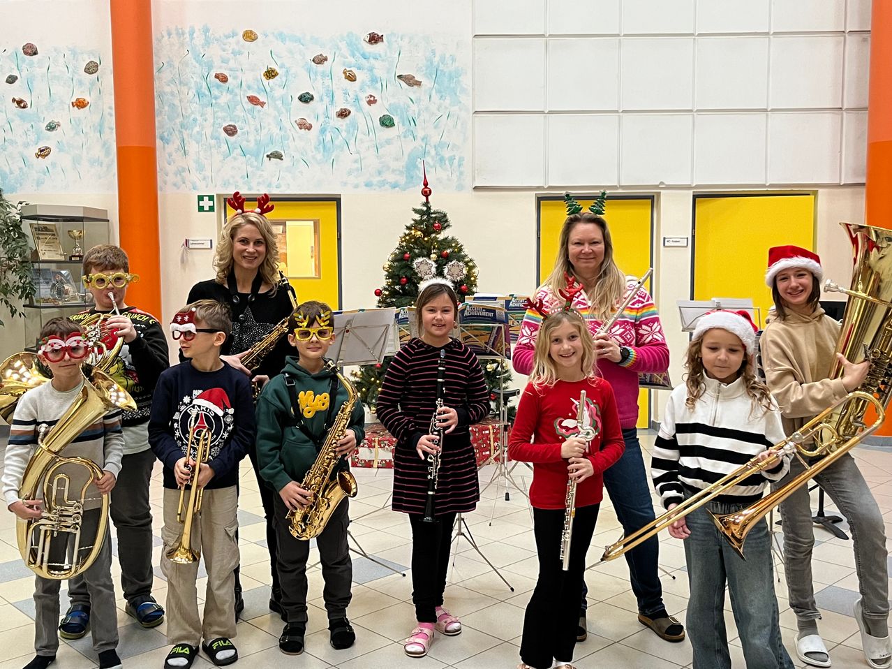 Eine Gruppe von Kindern und zwei Erwachsenen steht in einem Flur. Sie sind in weihnachtlich thematisierten Kleidungsstücken gekleidet und halten Blechblasinstrumente. Ein Weihnachtsbaum und Weihnachtsdekorationen sind im Hintergrund.