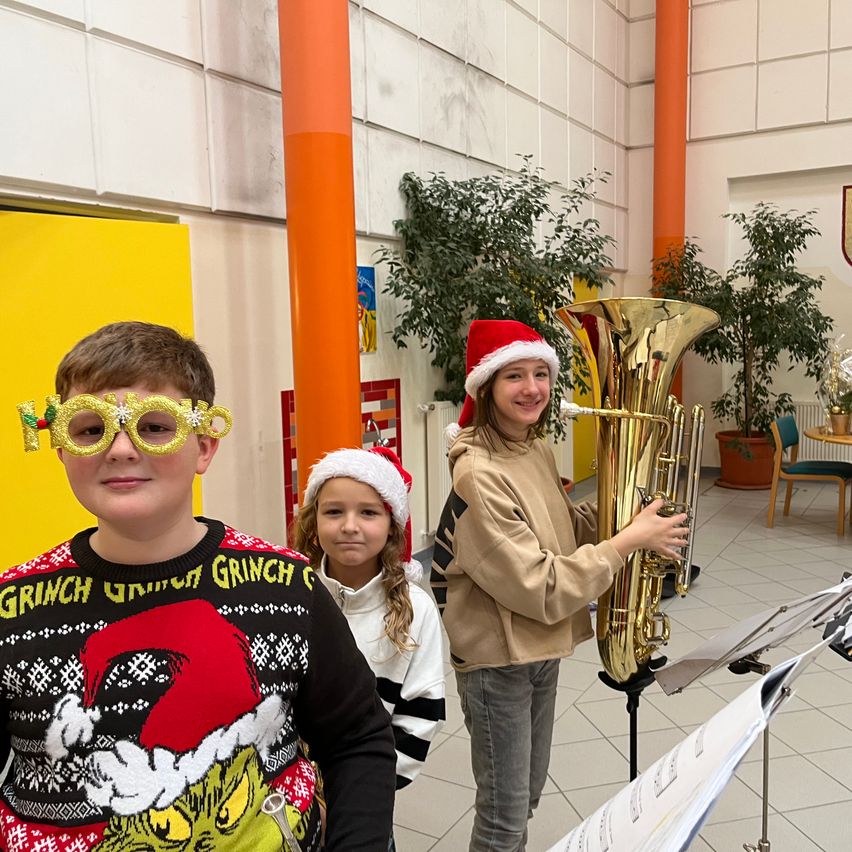 Drei Kinder sind für Weihnachten gekleidet. Ein Junge im Grinch-Pullover und mit Kulleraugen. Ein Mädchen mit Weihnachtsmütze neben ihm. Ein Mädchen mit Tuba hinter ihnen.