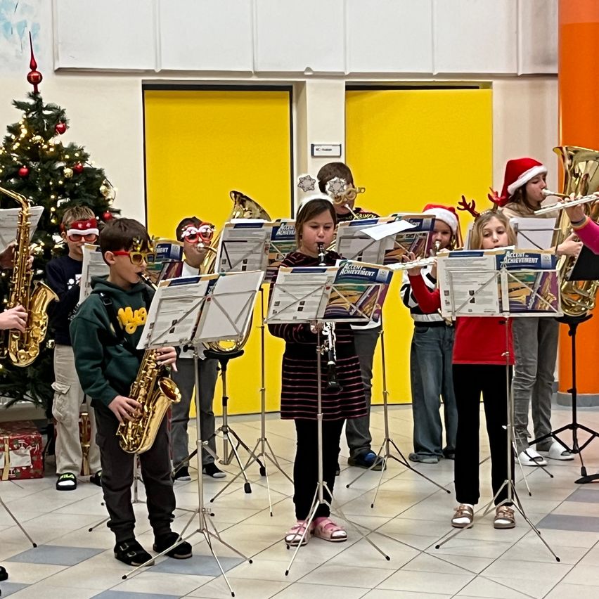 Eine Gruppe von Kindern in festlicher Kleidung, einschließlich Santa-Hüten, spielt Instrumente wie Saxophone und Trompeten in einem Flur. Ein Weihnachtsbaum ist im Hintergrund sichtbar.