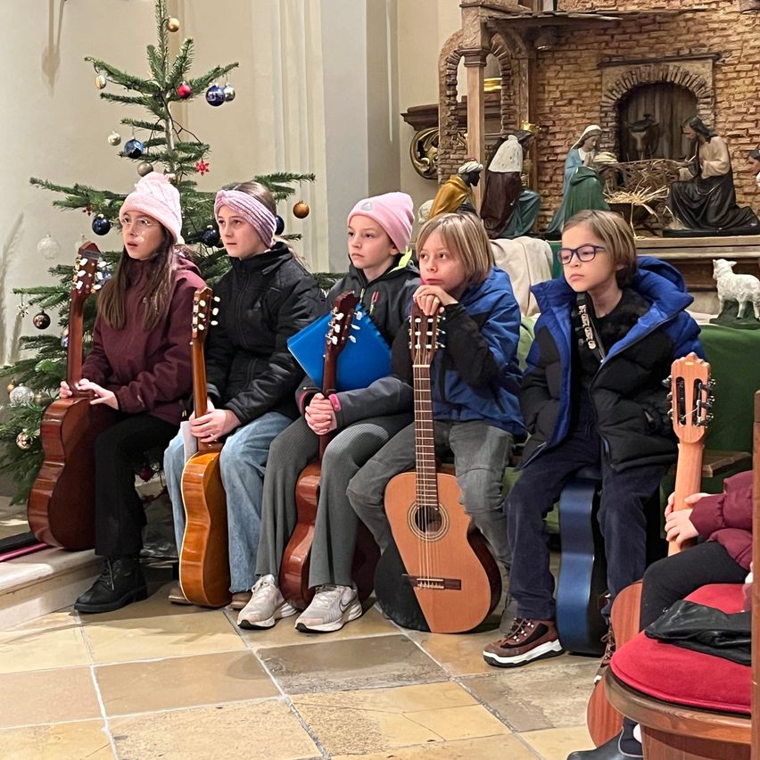 Fünf Kinder sitzen in einer Kirche und halten Gitarren. Ein Weihnachtsbaum und eine Krippe sind hinter ihnen. Sie sind warm gekleidet, was darauf hindeutet, dass es Winter ist.