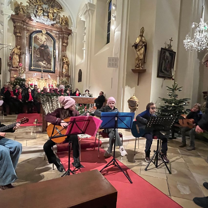 Eine Gruppe von Musikern spielt Gitarren in einer Kirche mit Weihnachtsdekorationen und Statuen an der Wand. Leute sitzen und stehen um sie herum.
