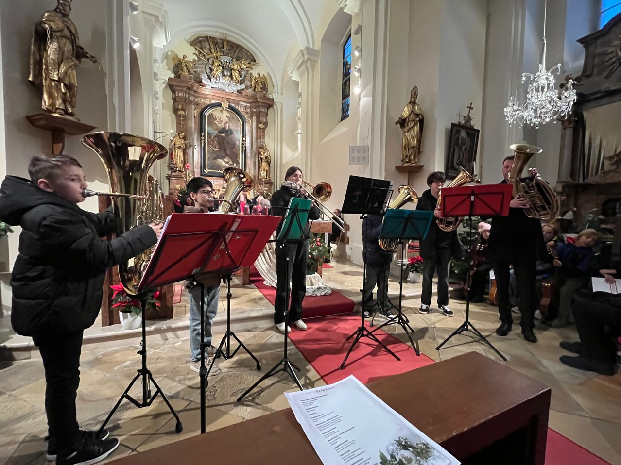 Eine Gruppe von Musikern spielt Blechblasinstrumente in einer Kirche. Die Musiker stehen auf einem roten Teppich vor einem Holzpult mit Musikblättern.