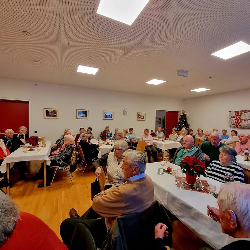 Eine große Gruppe älterer Menschen versammelt sich drinnen für eine Veranstaltung. Sie sitzen an Tischen mit Blumenarrangements und Gläsern. Der Raum hat weiße Wände, einen Holzboden und Weihnachtsdekorationen.