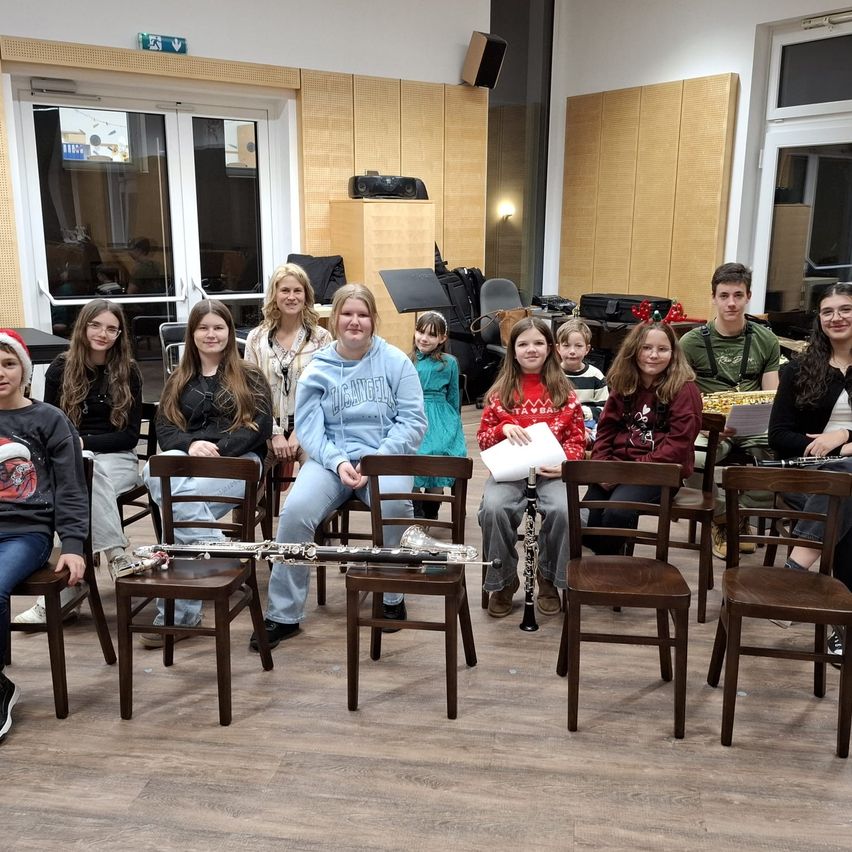 Eine Gruppe junger Menschen und Erwachsener sitzen mit Musikinstrumenten auf Stühlen, möglicherweise in einem Klassenzimmer oder Musikraum. Die Menschen lächeln und posieren für ein Foto. Zu den Instrumenten gehören Klarinette, Saxophon und Trompete. Der Raum hat Holzböden und -wände.