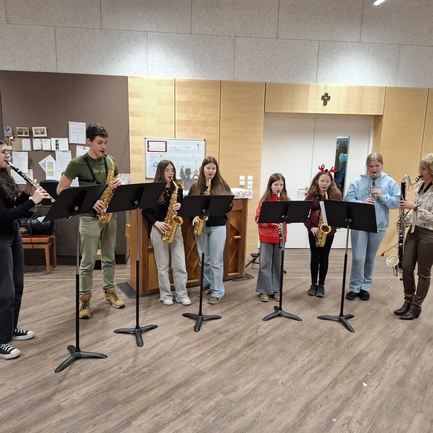 Eine Gruppe junger Musiker spielt in einem Raum mit Holzböden ihre Instrumente. Sie halten Saxophon und Klarinette. Hinter ihnen steht ein Holzschreibtisch mit Papieren darauf. Es gibt einen Holzstuhl und einen Schrank mit einem Kreuzsymbol oben.