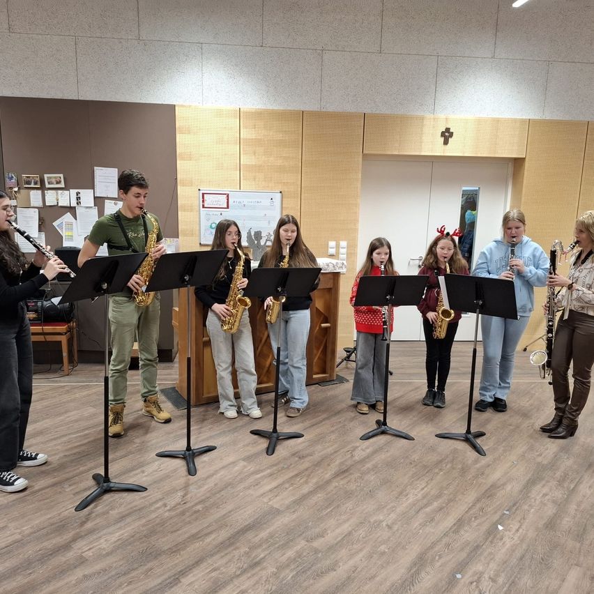 Eine Gruppe junger Musiker spielt in einem Raum mit Holzböden ihre Instrumente. Sie halten Saxophon und Klarinette. Hinter ihnen steht ein Holzschreibtisch mit Papieren darauf. Es gibt einen Holzstuhl und einen Schrank mit einem Kreuzsymbol oben.