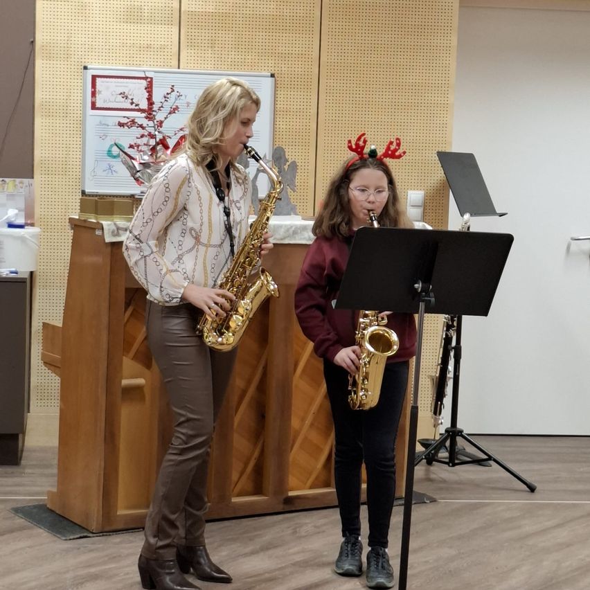 Eine Frau und ein Mädchen spielen Saxophon auf einer Bühne. Die Frau trägt ein weißes Oberteil und braune Hose, das Mädchen hat einen roten Haarreif. Ein Notenständer und ein Klavier stehen hinter ihnen.