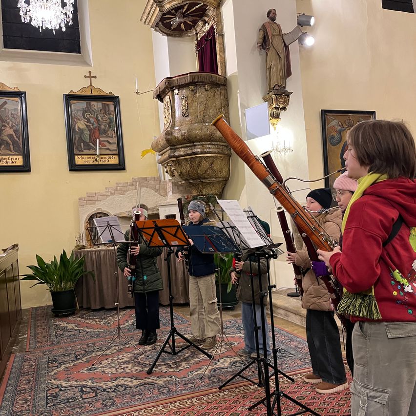 Eine Gruppe von Kindern spielt Blasinstrumente in einer Kirche. Die Instrumente umfassen Fagotte und Trompeten. Sie stehen auf einem Teppichboden und blicken zum Altar mit einer Statue darüber.