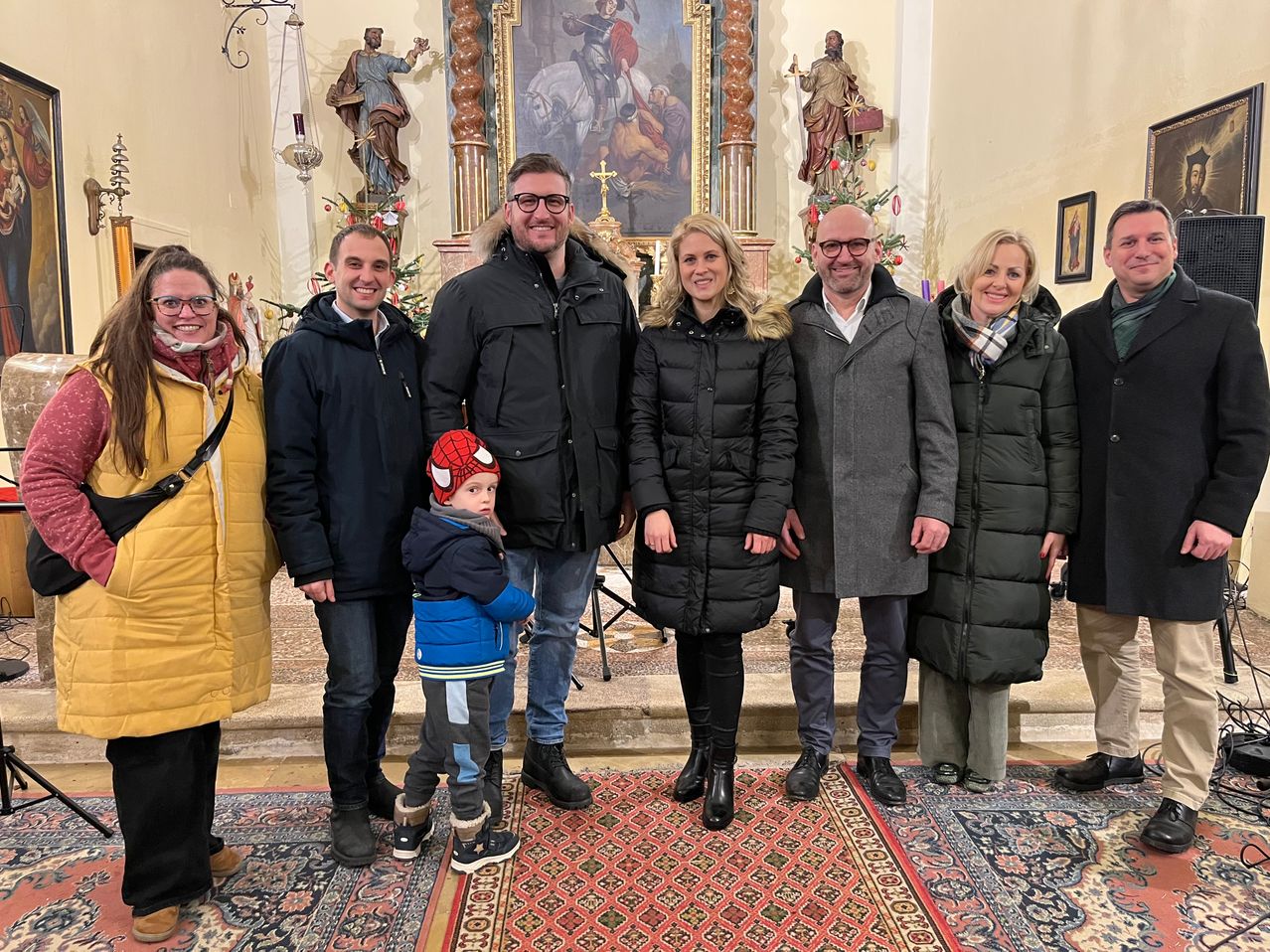 Eine Gruppe von Menschen, darunter ein Kind mit einer Spider-Man-Mütze, posiert für ein Foto in einer Kirche mit religiösen Statuen und einem Weihnachtsbaum im Hintergrund.