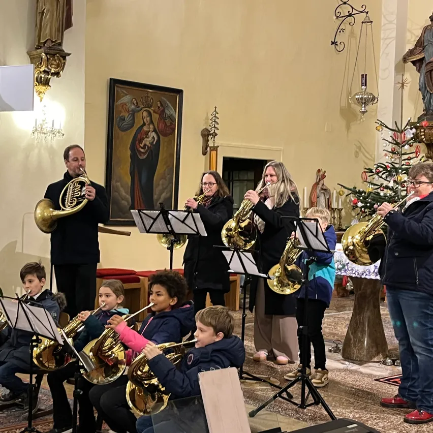 Eine Gruppe von Musikern spielt Blechblasinstrumente in einer Kirche, einschließlich Erwachsener und Kinder, mit einem Weihnachtsbaum im Hintergrund.