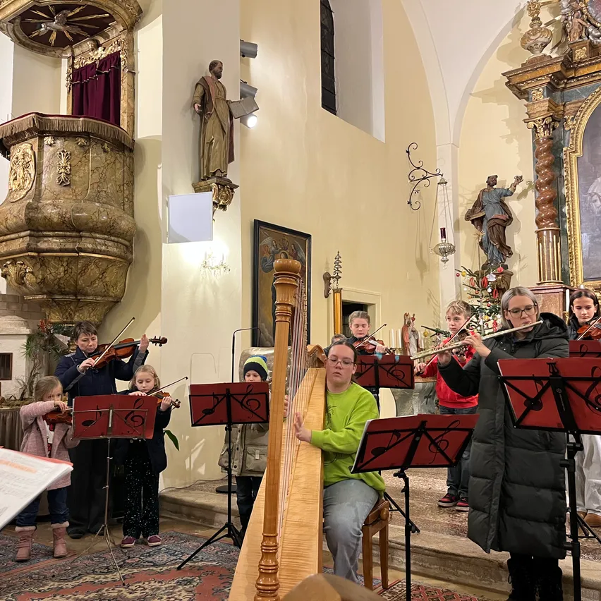 Eine Gruppe von Kindern spielt Musikinstrumente in einer Kirche, mit einem Harfenisten und einer Flötistin an der Spitze. Die Kinder stehen auf einem Teppich mit Notenständern.