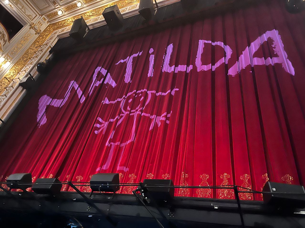 Ein roter Bühnenvorhang mit dem Wort Matilda in großen weißen Buchstaben und einer Silhouette eines Kindes. Die Bühne ist für eine Aufführung vorbereitet.