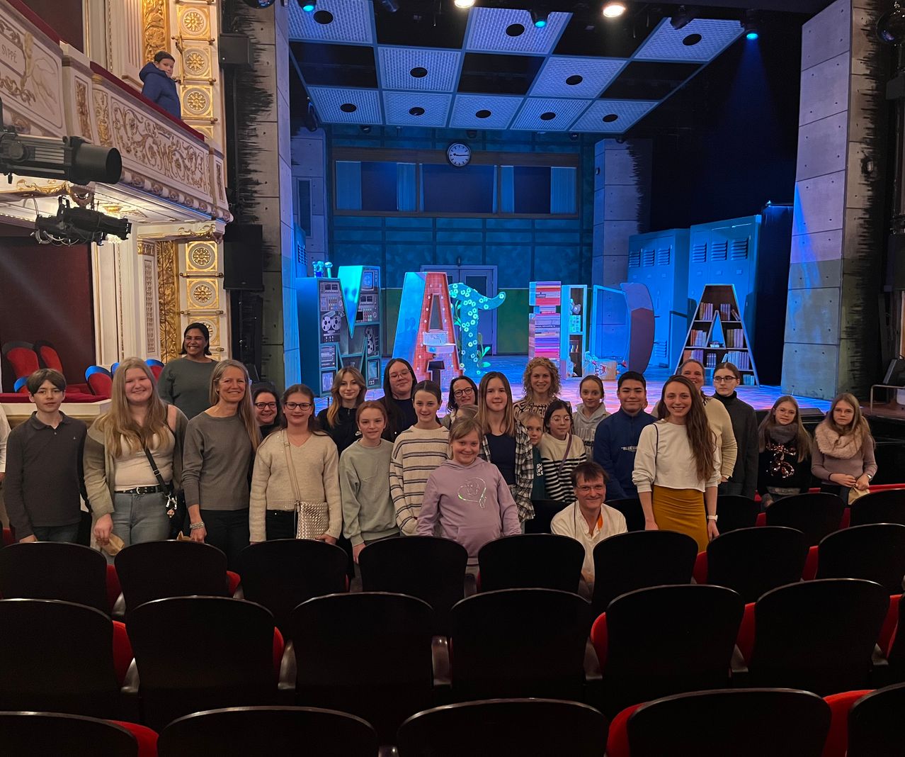 Eine Gruppe von Menschen in einem Theater posiert für ein Foto vor einer Bühne, die für ein Theaterstück vorbereitet wurde.