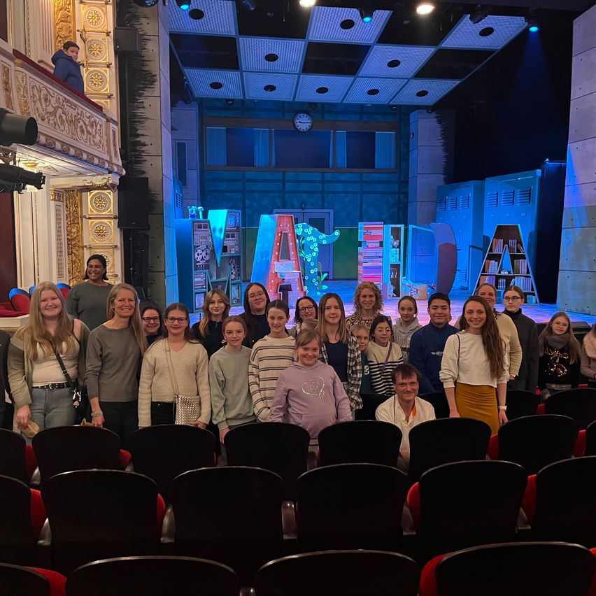 Eine Gruppe von Menschen in einem Theater posiert für ein Foto vor einer Bühne, die für ein Theaterstück vorbereitet wurde.