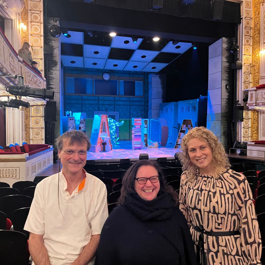 Drei Menschen lächeln vor einer Bühne mit bunten Lichtern und Requisiten. Hinter ihnen sind Reihen von Stühlen. Der Mann trägt ein weißes Hemd und eine Brille.