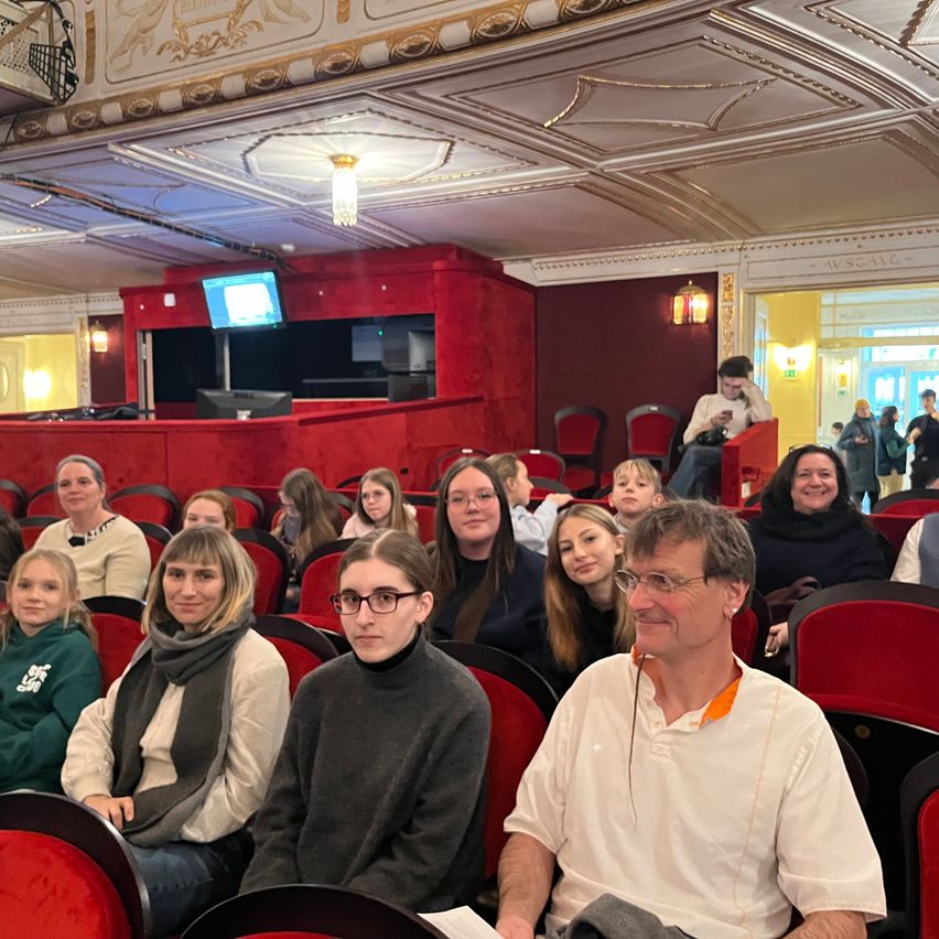Eine Gruppe von Menschen sitzt in einem Theater, möglicherweise bei einer Vorstellung. Einige tragen Brillen und Schals. Das Theater hat rote Sitze und dekorative Elemente. Eine Anzeigetafel befindet sich auf der Bühne.